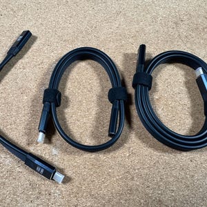 Puede incluir: Cables USB-C negros con correas de velcro negras. Los cables están enrollados y rectos, con conectores USB-C en cada extremo. Los cables están sobre una superficie marrón claro.