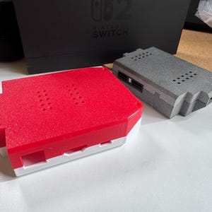 May include: Two 3D-printed cases, one red and white, the other grey, are on a white surface. The red case has a textured top with ventilation holes. A black Nintendo Switch box is in the background.