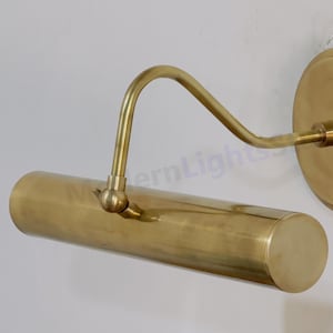 May include: A brass-colored wall-mounted picture light. The light has a cylindrical shade, a curved arm, and a round backplate. The fixture is designed to illuminate artwork or other wall-mounted objects.