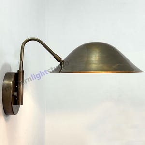 Peut inclure: Une applique murale en laiton antique avec un bras courbé et un abat-jour en forme de dôme. La lampe est fixée à une base circulaire et est conçue pour diriger la lumière vers le bas. Design minimaliste et industriel.