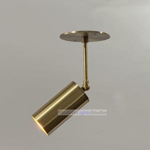 Puede incluir: Un foco ajustable de color latón. El cabezal cilíndrico está inclinado, con una luz brillante que emana de la abertura. La luminaria está fijada a una placa de techo redonda mediante una varilla recta.
