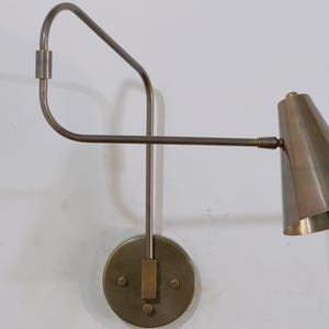 Puede incluir: Una lámpara de pared de latón con una pantalla cónica. La lámpara tiene un brazo curvo y una base circular con un detalle rectangular. El diseño es minimalista y moderno, adecuado para la iluminación interior.