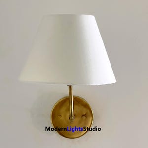 Puede incluir: Una lámpara de pared con una pantalla blanca y una base de color latón. La lámpara tiene una base circular y un brazo curvo que sostiene la pantalla. El texto "ModernLightsStudio" es visible en la base.