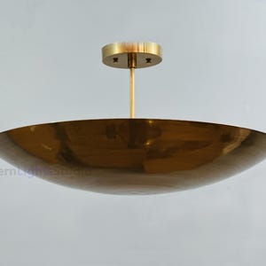 May include: A contemporary ceiling light in a brass finish. It features a shallow, bowl-shaped shade hanging from a brass rod and a circular ceiling fitting. The design is minimalist and stylish, suitable for various interiors.