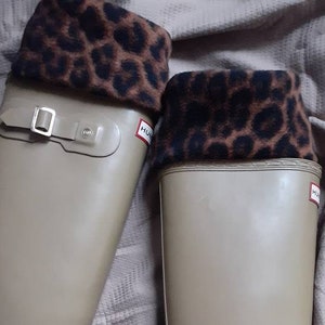 May include: Two tall, tan wellington boots with a matte finish. Each boot features a buckle detail and a brown and black leopard print cuff. The boots are set against a neutral-toned fabric background.