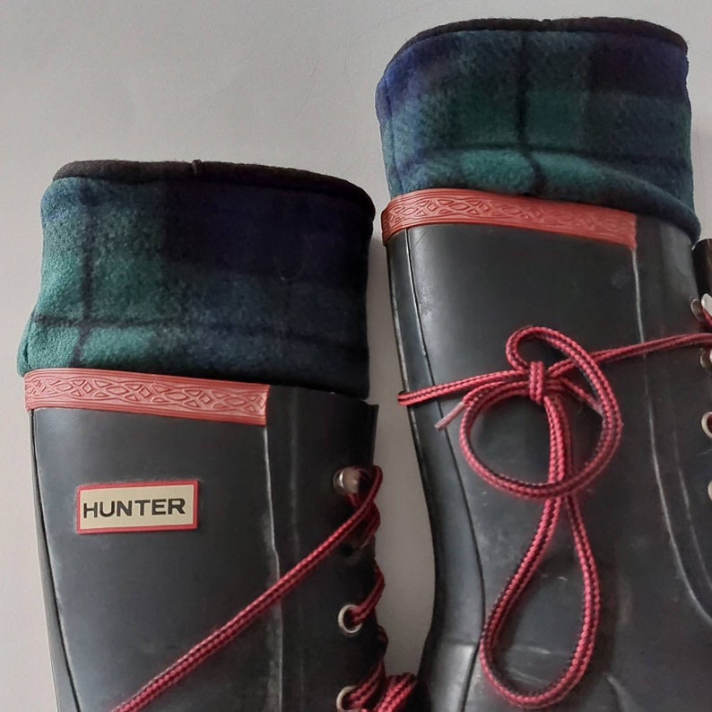 May include: Pair of black Hunter wellington boots with red laces and a green, blue, and black plaid fleece boot cuff. The boots have red trim and the word "HUNTER".