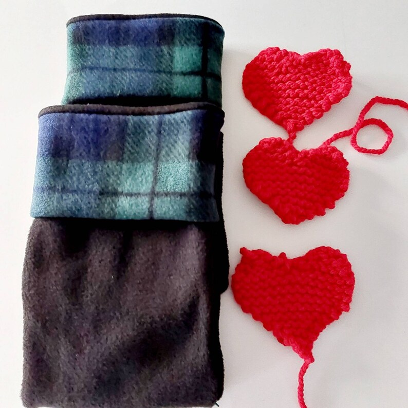 May include: Three fleece neck warmers in black, green, and blue plaid. Three red knitted hearts are also displayed. The neck warmers are folded and stacked. The hearts are connected by yarn.