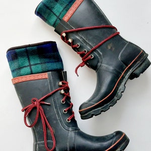 May include: A pair of black wellington boots with red laces and a green and blue tartan cuff. The boots have a red trim and metal eyelets. The boots are designed for outdoor wear.
