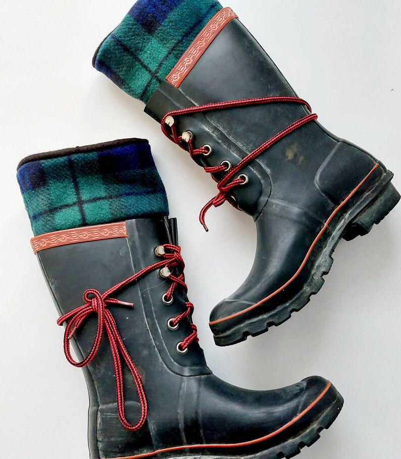 May include: A pair of black wellington boots with red laces and a green and blue tartan cuff. The boots have a red trim and metal eyelets. The boots are designed for outdoor wear.