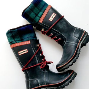 May include: Pair of black Hunter wellington boots with red laces and trim. The boots feature a green, blue, and black plaid fabric cuff. The word "HUNTER" is on a red rectangle on the side of each boot.