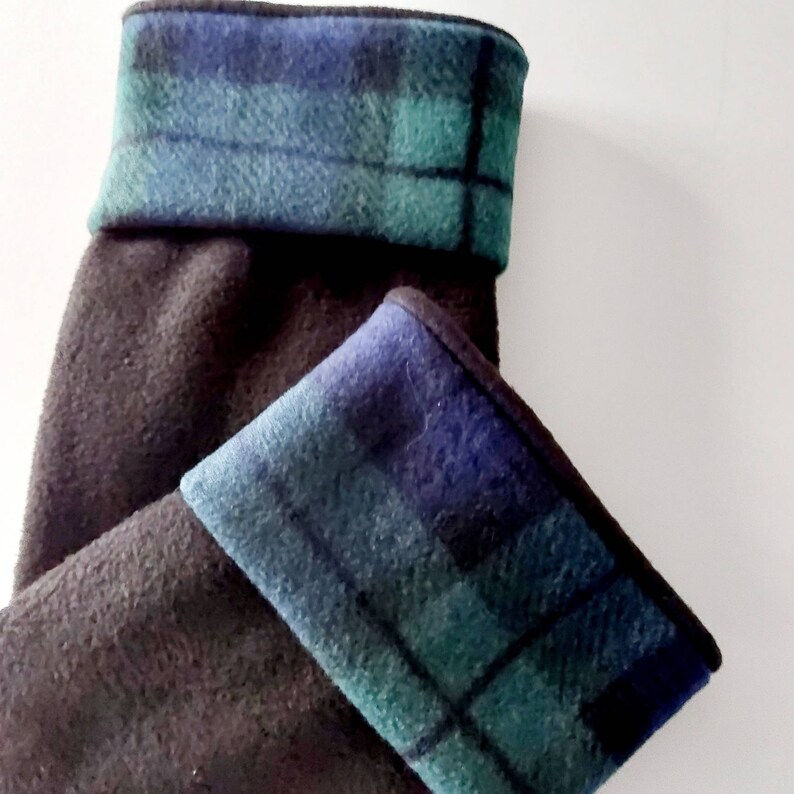 May include: Dark grey fleece leg warmers with a folded cuff. The cuffs display a blue and green tartan pattern. These leg warmers are designed to keep legs warm and are suitable for various activities.
