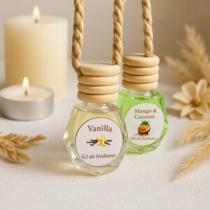 May include: Two hanging air fresheners with wooden tops and hexagonal glass bottles. One is labelled "Vanilla" with a flower graphic, the other "Mango & Coconut." Both have beige rope hangers. Lit candles and dried flowers are in the background.