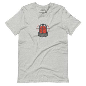 May include: A light gray t-shirt featuring a cartoon illustration of a red emergency light with a gray base and cage. The shirt has a crew neck and short sleeves. The shirt is made of a soft, comfortable material.