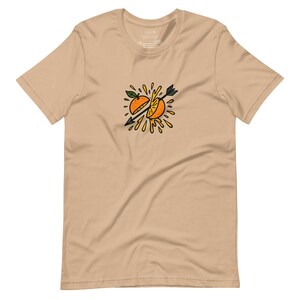 May include: Tan short-sleeve t-shirt featuring a graphic of an orange sliced in half by an arrow. The orange is yellow and orange with green leaves, and the arrow is black. The design is centered on the chest.