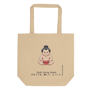 Puede incluir: Bolso tote beige con una ilustración de dibujos animados de un luchador de sumo. El luchador tiene una expresión seria y lleva un mawashi rojo. El bolso presenta el texto "Small, strong, steady" en inglés y japonés.