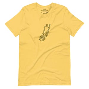 Puede incluir: Camiseta amarilla con un dibujo de contorno negro de un teléfono plegable. La camiseta tiene cuello redondo y mangas cortas. El texto de la etiqueta dice "Sustainable is the new sexy".