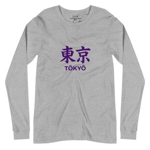 Könnte beinhalten: Ein langärmeliges T-Shirt in Grau meliert mit den violetten japanischen Kanji-Schriftzeichen für Tokio über dem Text TŌKYŌ in einer fetten, serifenlosen Schrift. Das Shirt hat einen Rundhalsausschnitt und lange Ärmel mit gerippten Bündchen.