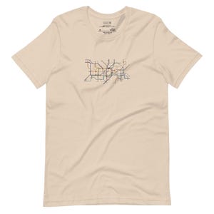 May include: A cream-colored t-shirt featuring a colorful subway map design. The map is a network of lines in various colors, including blue, orange, and brown. The shirt has a classic crew neck and short sleeves.