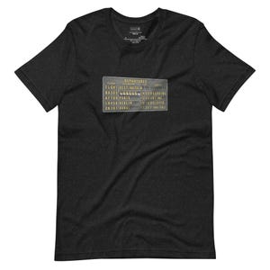 Puede incluir: Camiseta gris oscuro con un diseño gráfico de tablero de salidas de estilo vintage. El tablero muestra destinos como París y Dubái, con horarios y estados de los vuelos. Una camiseta para los amantes de los viajes.