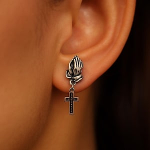 May include: A silver-toned earring featuring praying hands above a dangling cross. The cross has a detailed design with small dots. The earring is worn on an ear, with the focus on the religious-themed jewelry.