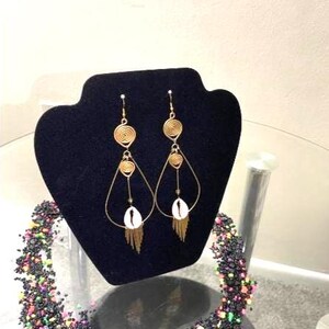 May include: Gold-toned earrings with spiral and teardrop designs, featuring a white shell and dangling fringe. The earrings are displayed on a black velvet stand. A beaded necklace with colorful accents is in the foreground.