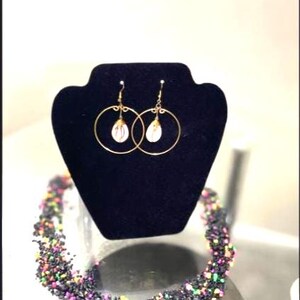 May include: Gold hoop earrings with teardrop-shaped white and pink accents are displayed on a black velvet stand. A beaded necklace with black, purple, green, and pink beads is in the foreground.