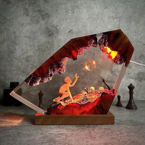 May include: A unique resin lamp featuring a fantasy scene with a figure reaching for a glowing ring. The lamp has a wooden base and a clear, angled frame with red and orange accents, and a light inside.