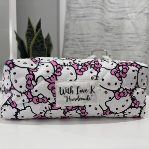 Puede incluir: Un neceser rectangular blanco con un estampado repetido de caras de Hello Kitty y lazos rosas. La bolsa tiene una pequeña etiqueta que dice "With Love K Handmade".