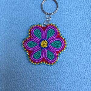 May include: A beaded flower keyring with a red centre, purple petals, and teal accents. The flower is outlined with yellow and blue beads, attached to a silver keyring. The background is a light blue textured surface.
