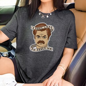 May include: A dark charcoal grey short-sleeve t-shirt featuring a graphic illustration of a man with a moustache. The design includes a banner with the text I KNOW WHAT I'M ABOUT SON wrapped around the illustration in a light beige colour.