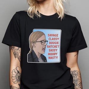 May include: A black short-sleeved t-shirt featuring a graphic print of a person with blonde hair and glasses, mouth open in an expression. Next to the figure, the text reads: SAVAGE, CLASSY, BOUGIE, RATCHET, SASSY, MOODY, NASTY in red capital letters.