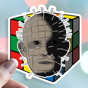 May include: A sticker featuring a black and white illustration of a character with a head covered in pins, set against a colorful Rubik's Cube background.