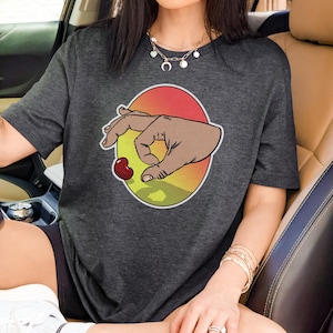 May include: A charcoal gray short-sleeve t-shirt featuring a graphic print of a hand making a pinching gesture toward a small red cherry. The design is set against a circular background with a gradient transitioning from orange to yellow to green.