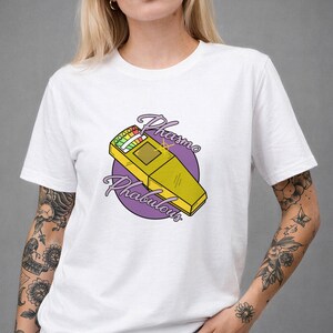 May include: A white short-sleeve t-shirt featuring a graphic of a yellow EMF meter on a purple circular background. The text "Phasmo Phabulous" is written in a purple script font curving around the yellow device, which has a small light display.