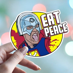 May include: A sticker featuring a cartoon character with a blue and red costume, a silver helmet, and a tongue sticking out. The character is surrounded by yellow and red splatters. The text "EAT PEACE" is written in white letters on a red background.