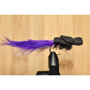 May include: A fishing fly with a black body and a bright purple tail. The fly is attached to a hook and is designed to resemble an insect. This fly is likely used for fly fishing.