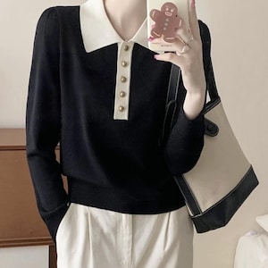 May include: Black long-sleeve sweater with a white collar and gold buttons. The sweater has a polo-style neckline and is paired with white pants. A beige and black tote bag is also visible.