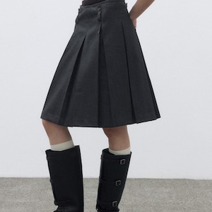 May include: A dark gray pleated skirt with a button closure, paired with black knee-high boots. The boots have multiple buckles and a lace-up design. The outfit is completed with a black top.