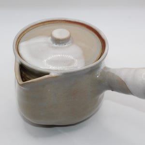 Puede incluir: Una tetera de cerámica con tapa y asa. La tetera es principalmente de color marrón claro con un asa y tapa blancas. El pico es triangular. La tetera es probablemente hecha a mano.