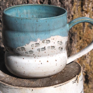 May include: A ceramic mug with a rounded, bulbous shape featuring a two-tone glaze. The upper portion is a smooth, glossy turquoise blue, while the lower section is a speckled white with small, dark brown boot print patterns across the surface.