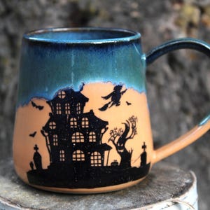 Peut inclure: Mug en céramique sur le thème d'Halloween avec une silhouette de maison hantée noire. Le mug a un dessus bleu et vert, un milieu orange et une anse noire. Le design comprend une sorcière sur un balai.
