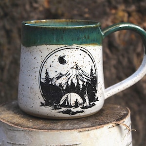 Op de afbeelding: Een gespikkelde keramische mok met een groene rand en handvat. De mok heeft een zwart-wit illustratie van een berglandschap met een tent, bomen en een maan. De mok staat op een houten oppervlak.