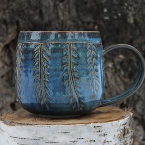 Puede incluir: Una taza de cerámica azul hecha a mano con un diseño de hojas. La taza tiene una forma redondeada con un asa y está sobre una superficie de madera. El diseño de la taza presenta una serie de patrones verticales en forma de hojas.