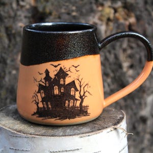 May include: A Halloween-themed ceramic mug. It has a black rim and handle, and an orange body. The front is decorated with a spooky haunted house design and flying bats. The mug is placed on a wooden surface.