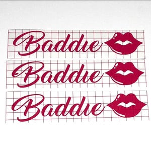 May include: Three red vinyl decals with the word "Baddie" in a cursive font, each accompanied by a red lip graphic. The decals are on a grid background, suitable for crafting or personalization projects.