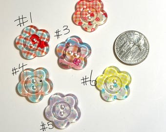 Pode incluir: Uma coleção de seis botões em forma de flor em várias cores e padrões. Os botões têm um acabamento transparente e brilhante e apresentam diferentes designs, incluindo xadrez e estampas florais. Uma moeda de 25 centavos de dólar americano está incluída para referência de tamanho.