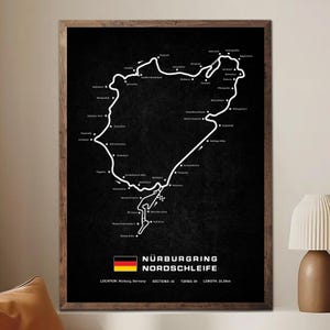 Puede incluir: Un grabado en blanco y negro enmarcado del circuito de carreras Nürburgring Nordschleife. El diseño de la pista está representado en blanco sobre un fondo negro. El grabado incluye la bandera alemana y el texto: Nürburgring Nordschleife, Secciones: 44, Curvas: 89, Longitud: 25,3 km.