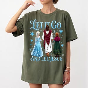 May include: Olive green t-shirt featuring the text "Let It Go and Let Jesus" in blue script. The graphic depicts Jesus holding hands with two figures in blue and green dresses, with snowflakes.