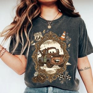K&ouml;nnte beinhalten: Dunkelgraues T-Shirt mit einem Cartoon-Bild von Hook aus dem Film Cars, in einem verzierten Rahmen mit Route 66-Text. Das Design umfasst einen Verkehrskegel, Blitze und Zielflaggen.