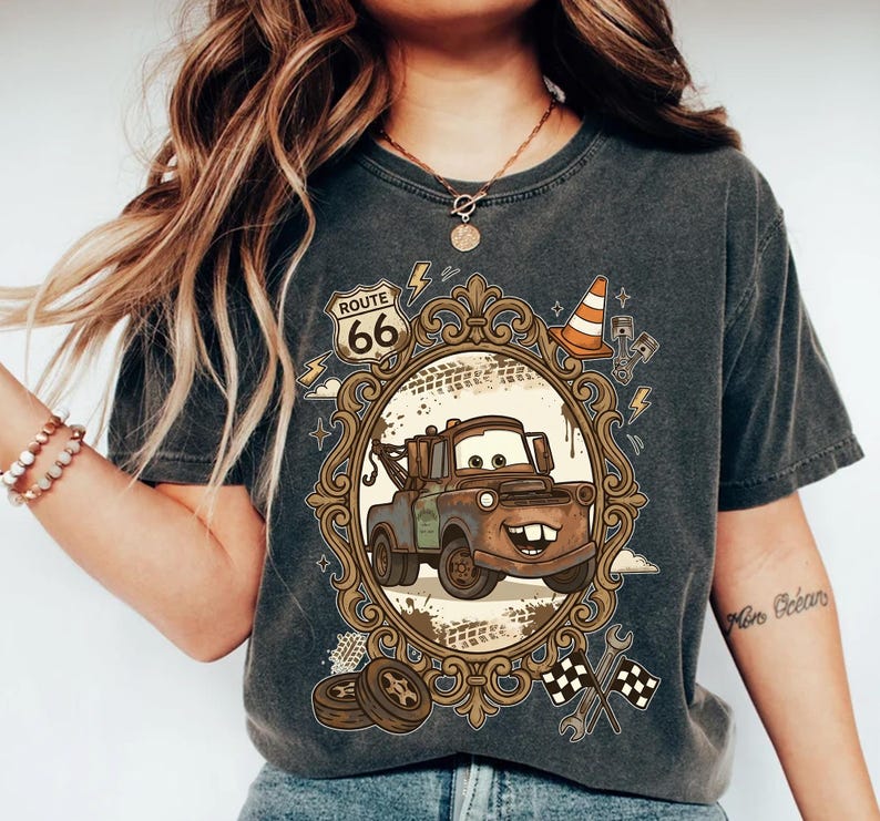 K&ouml;nnte beinhalten: Dunkelgraues T-Shirt mit einem Cartoon-Bild von Hook aus dem Film Cars, in einem verzierten Rahmen mit Route 66-Text. Das Design umfasst einen Verkehrskegel, Blitze und Zielflaggen.